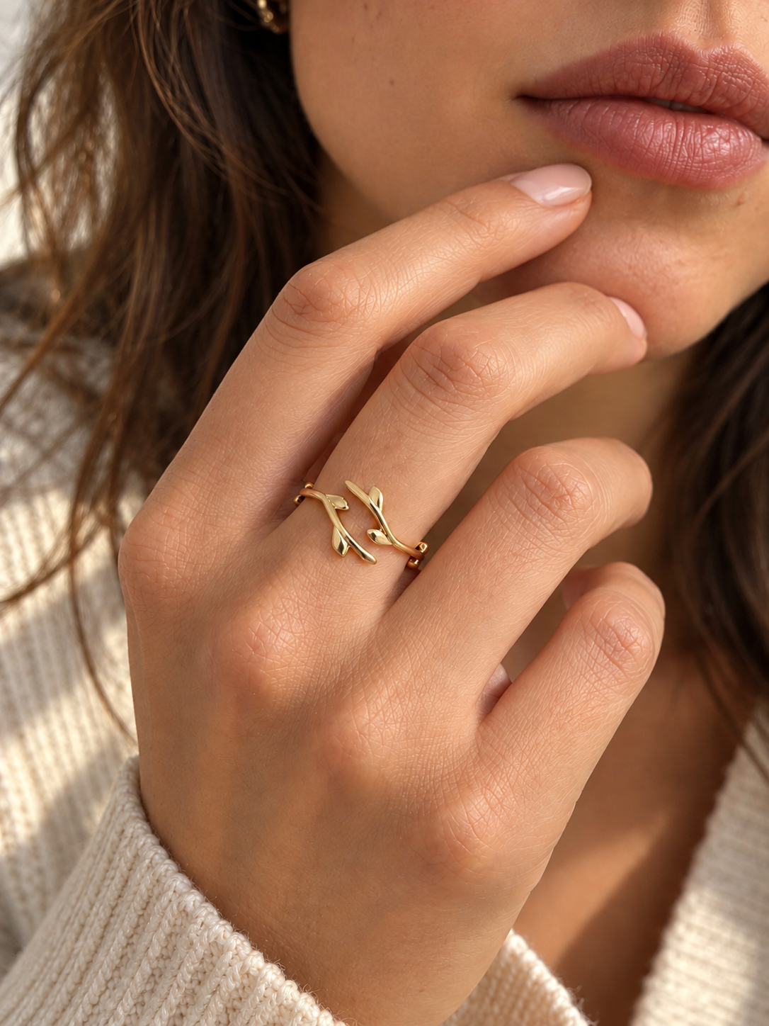 A close-up of a woman's hand wearing a delicate gold ring designed to look like olive or willow branches or leaves, styled with a cream knit sweater.